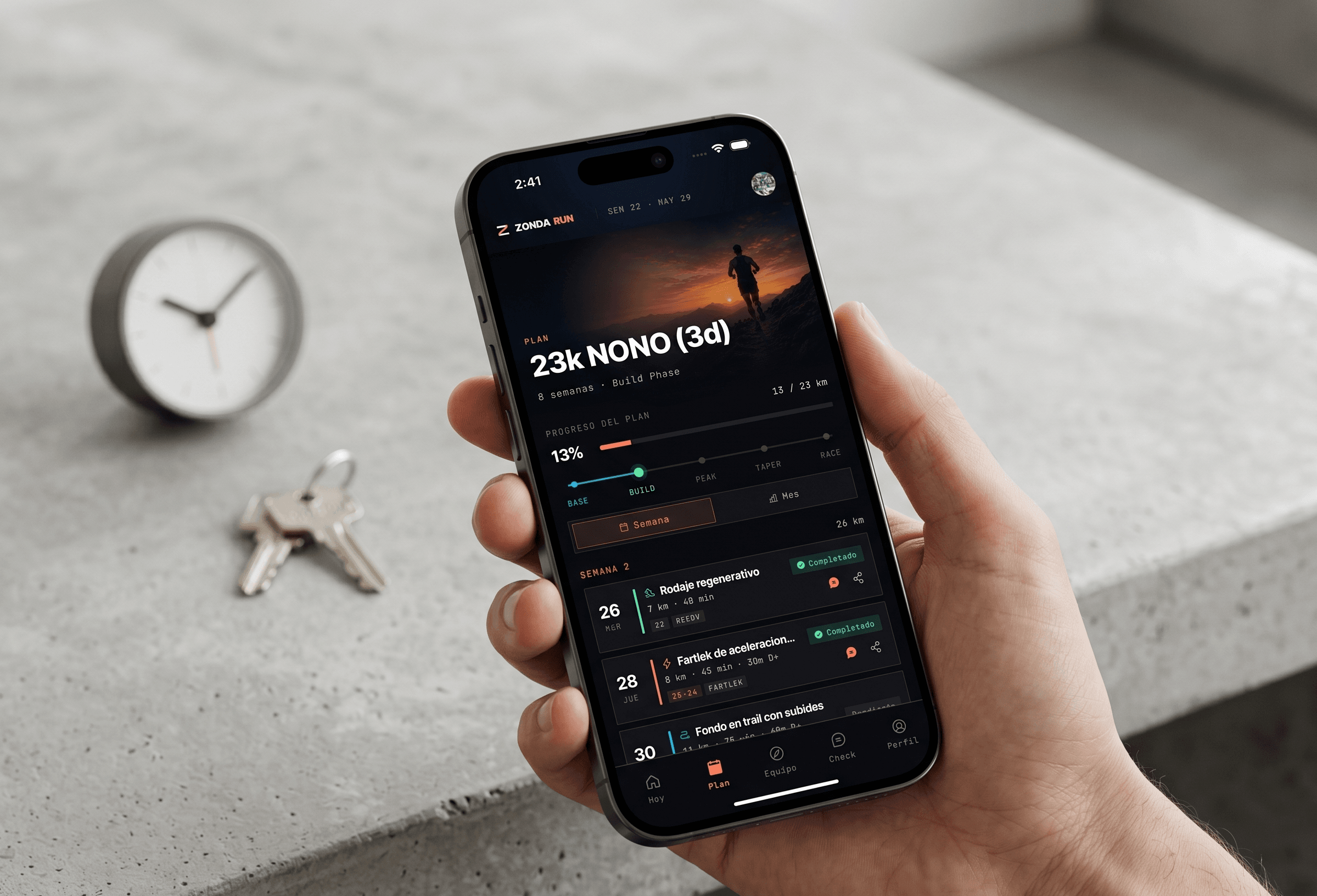 Mano sosteniendo un iPhone con la app de Zonda Run abierta en el plan '23k NONO' del atleta: calendario semanal con sesiones (Rodaje regenerativo, Fartlek de aceleración, Fondo en trail con subidas) y métricas de progreso, sobre fondo de concreto con llaves y reloj.
